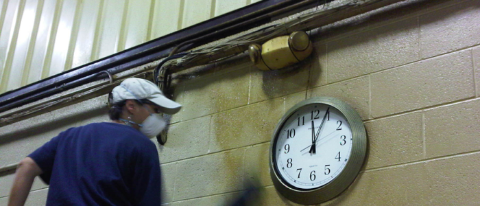 Woman wearing a blue shirt, beige ball cap with a white face mask and blue gloves cleaning a greasy concrete block wall area next to a large wall clock showing time of 12:05
