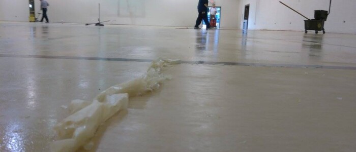 Close up photo of scraped and accumulated floor finish in an empty church gym with 3 floor techs and mop bucket and wet vac equipment in the backgroud.