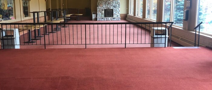 Clean red colored carpet in a bi-level large empty dining area with big windows on the right and a stone fireplace in the back with stacked tables and 2 garbage cans.